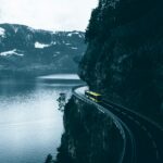 Żółty autobus jadący krętą drogą wykutą w skale nad brzegiem jeziora w górzystym terenie.